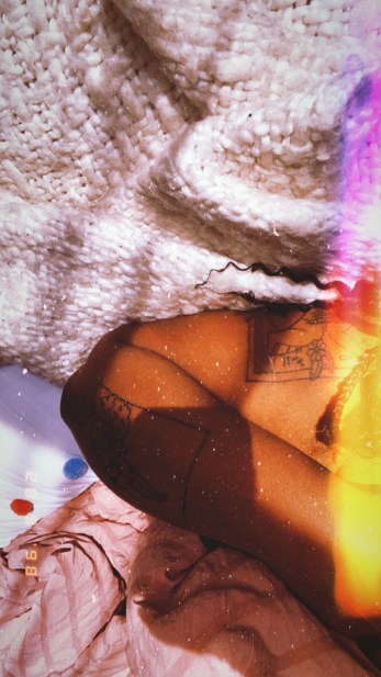 Image shows black woman in bed with tattoos on her arm with 3 different textured blankets surrounding her.