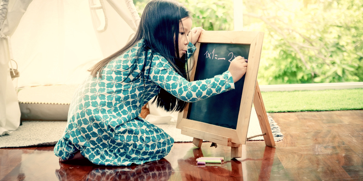 A young girl is in a green dress and bent off a chalkboard as she works on a math problem against a spring background, it looks serene.