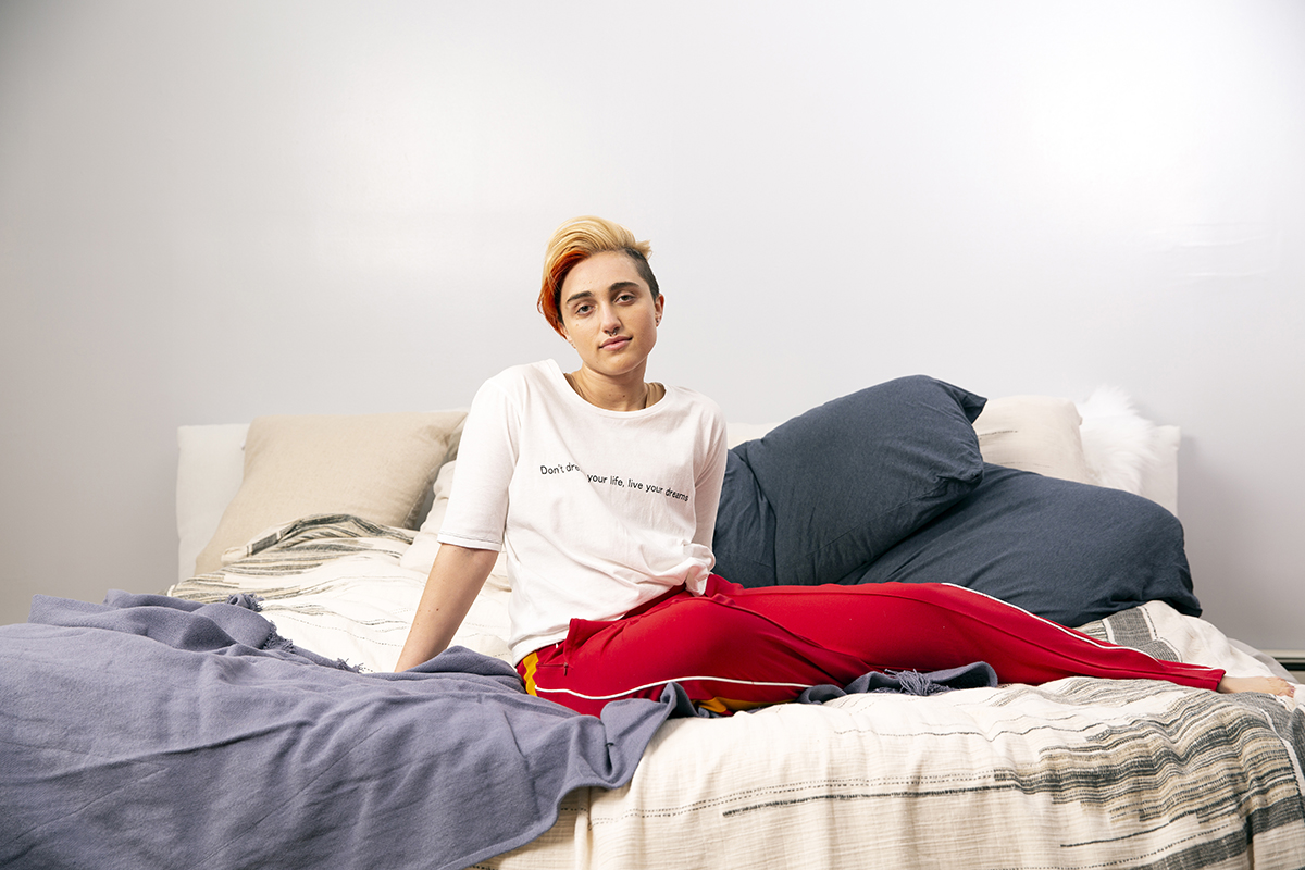 A transmasculine gender-nonconforming person sitting on a bed.