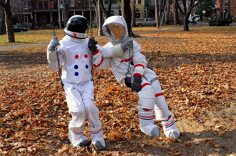 Two figures in spacesuits sit on a swingset in a sunny park full of fall leaves, one leaning toward the other as if to embrace them