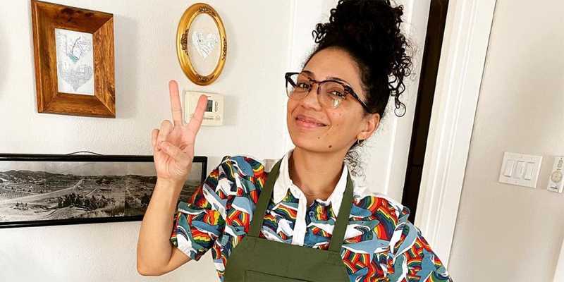 Jasika Nicole has her curly hair up in a top knot bun and she is wearing glasses. She has on a multicolored t shirt with a dark green apron and is flashing a peace sign at the camera.