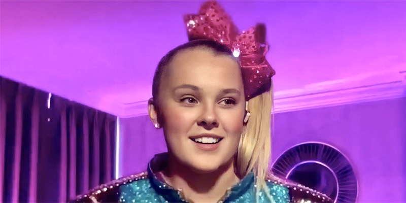 JoJo Siwa has a bright pink bow and turquoise sparkly jacket. She's against a purple background in her room. Her hair is in her famous side ponytail. She stares confidently to her left as she ponders how great it is to be gay.