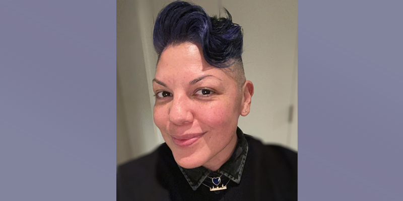 A selfie of Sara Ramirez with their signature hair cut died a dark lavender purple. They are smirking at the camera, full of pride and mischief.