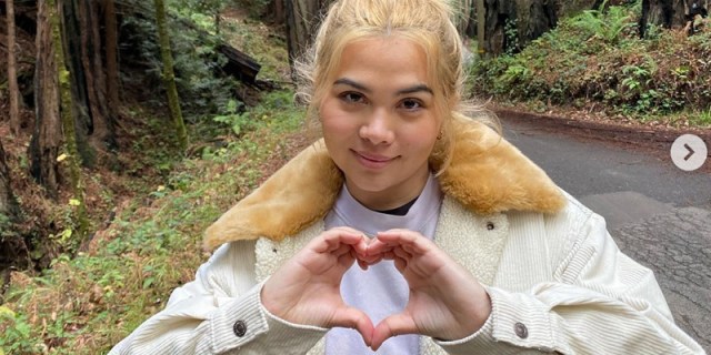 hayley kiyoko is in the forest and makes a heart shape with her hands. She is in a white winter coat with blonde hair in a bun.