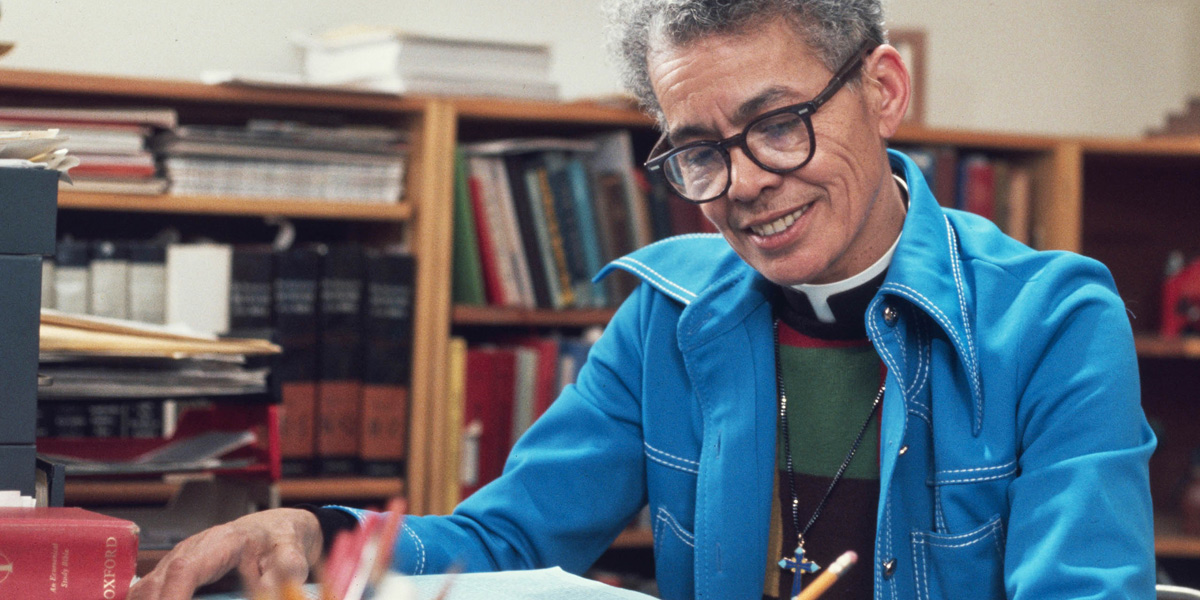 Pauli Murray wears glasses and short cut grey hair with a denim blue overskirt while smiling and writing in a library in front of books.