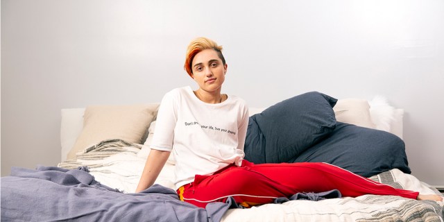 A transmasculine gender-nonconforming person sitting on a bed.