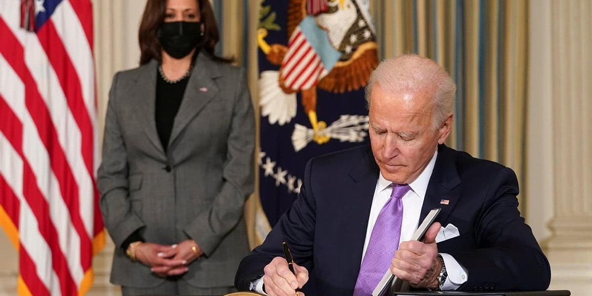 President Joe Biden signs papers in the White House with Vice President Kamala Harris in the background.