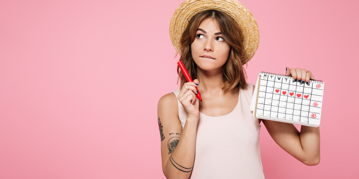 A tattooed woman with brown hair in a straw hat stands in front of a pink background. She holds a red pen and a calendar that has some days marked with red hearts.