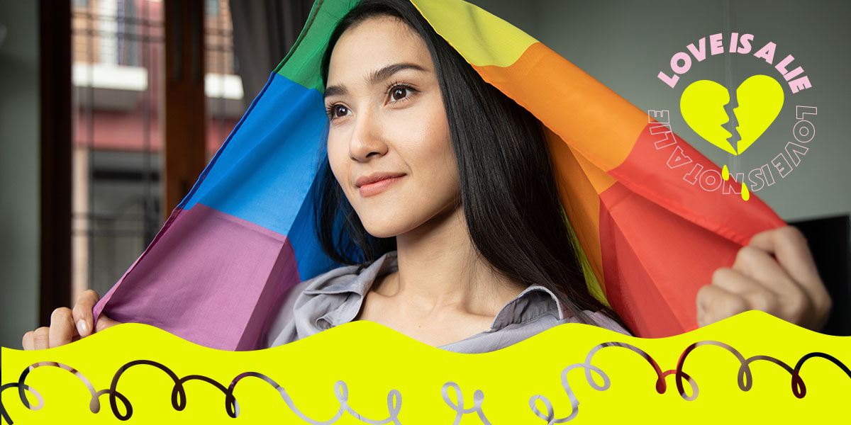 A person stands with a rainbow flag draped over their head, staring off into the middle distance.