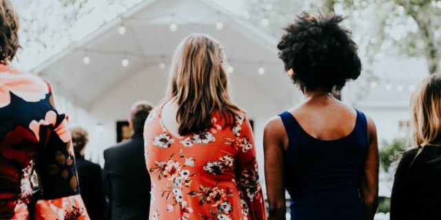 Two young women photographed from behind in the audience of a public outdoor event, one white and wearing a red floral print dress with shoulder-length brown hair, and one Black and wearing a black tank top with natural hair