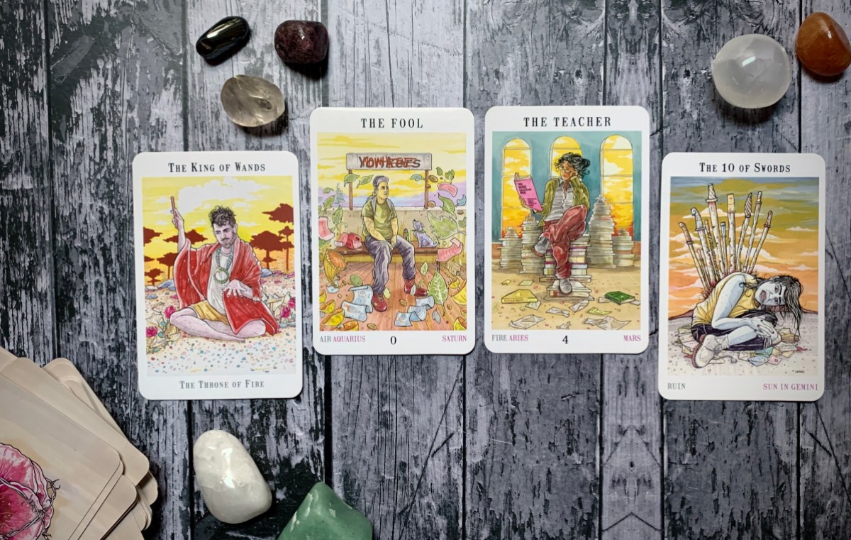 Four tarot cards from the Next World deck arranged on a wooden surface surrounded by some stones and crystals; from left to right, the cards are the King of Wands, the Fool, the Teacher and the Ten of Swords.