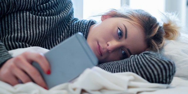 A troubled-looking young woman lies on her side in a bed with her phone in front of her face, looking past it into the distance pensively