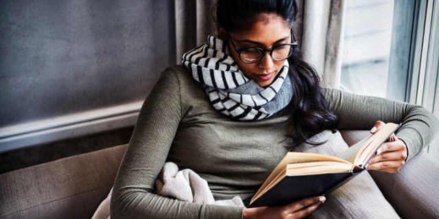 A person with long dark hair in a ponytail wearing glasses and a scarf reads a book curled up in a window corner.