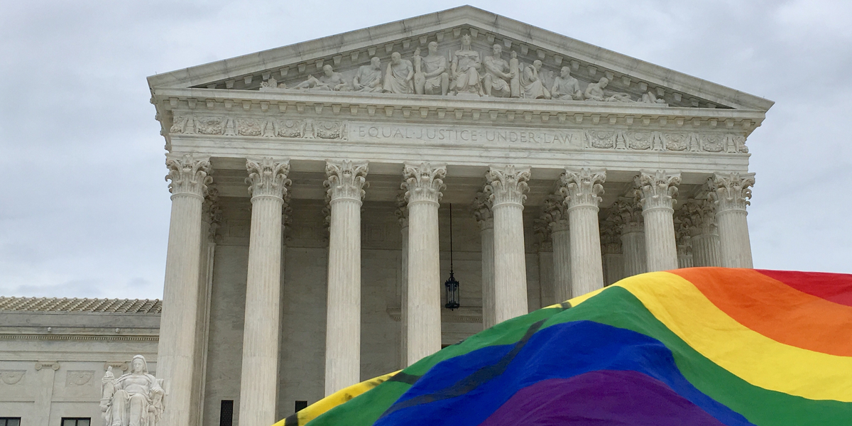 A government building photographed from afar, with the corner of a rainbow flag fluttering in the foreground.