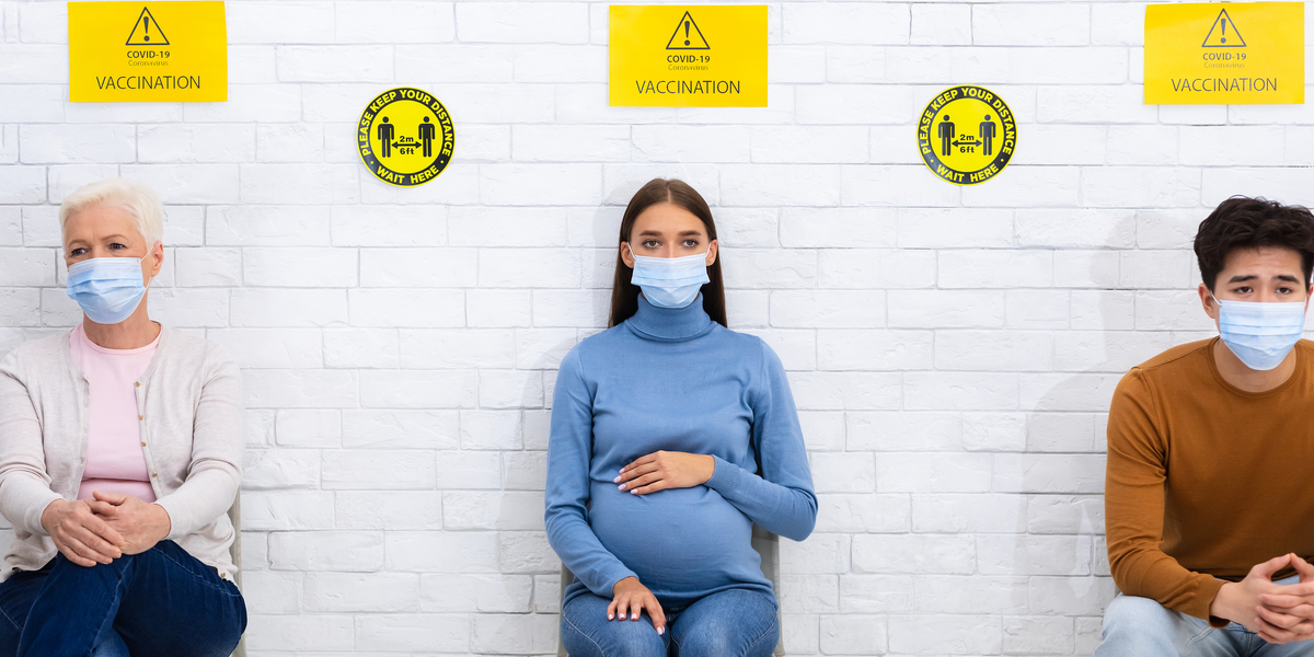 Three people sit in socially distanced chairs, wearing masks as signs above their heads indicate they're in line to receive the COVID-19 vaccine.