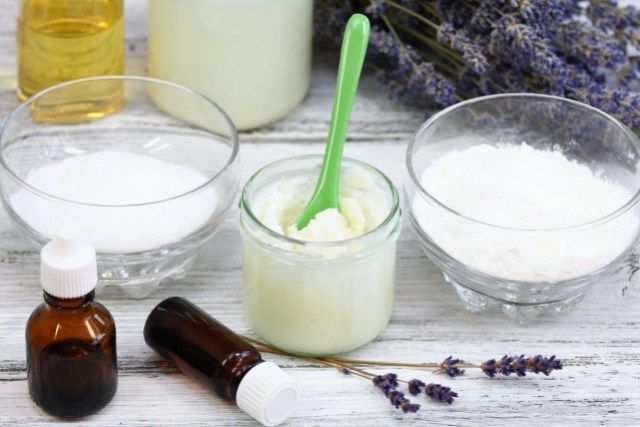 Two bowls containing powder and one jar containing oil are on a table near essentials oils and sprigs of lavender