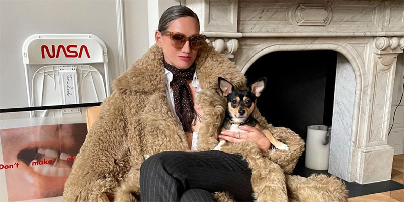 Style Maven Jenna Lyons relaxes in a full length faux fur coat and her hair slicked back in a bun and dark cateye sunglasses. She is holding a small dog in her arms and is in front of a fire place. She's a boss, personified.