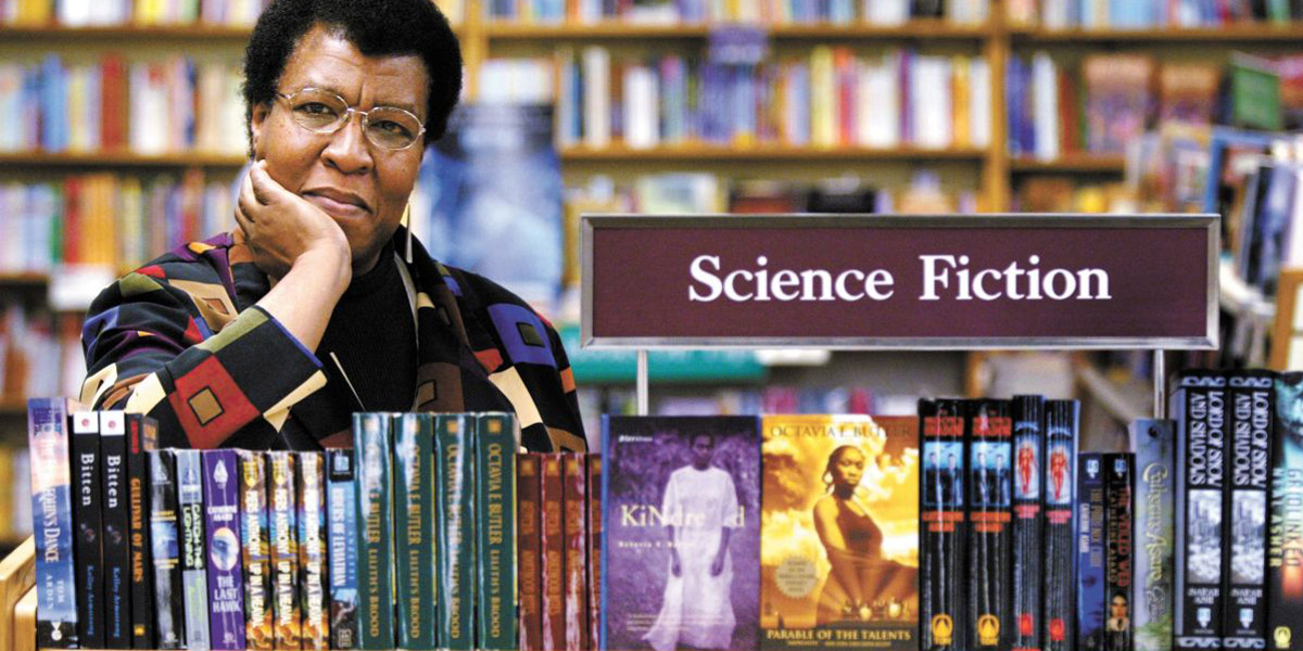 Octavia Butler in the 1990s stands in front of a collection of her books under the sign "Science Fiction" in a library