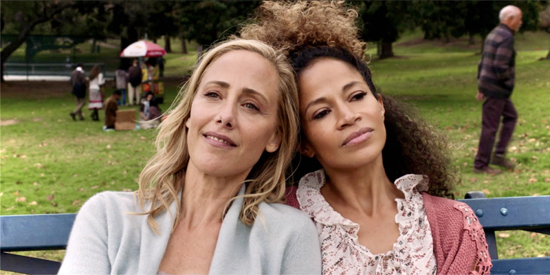 Dr. Teddy Altman sits on a bench with her ex-girlfriend in Central Park. The bench is blue, the grass is green, and the girlfriends rest their heads on each other's shoulders in love.