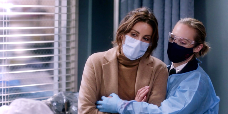 In a still from Grey's Anatomy, Carina Deluca is wearing a mask and being held by her girlfriend Maya Bishop, who is in scrubs and also a mask. They both look distraught and worried as they stare off to the left of the camera.