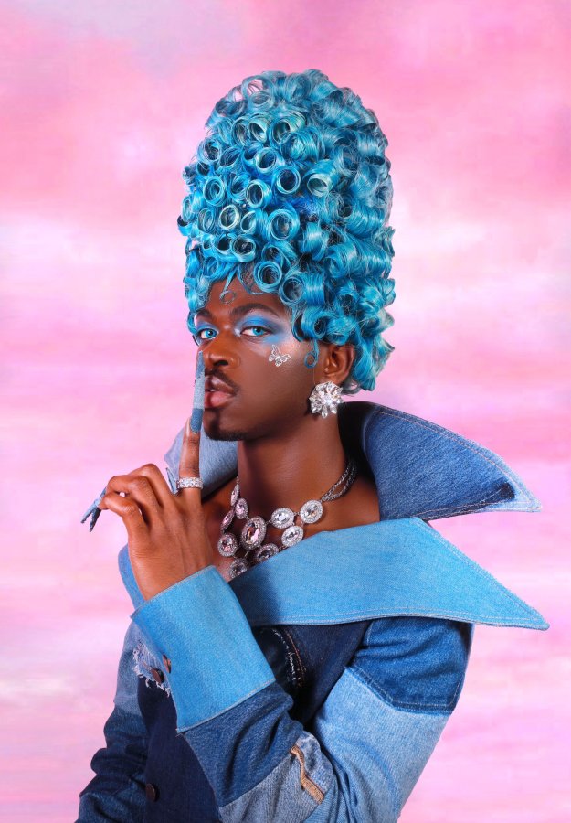Lil Nas X queer promo Image shows Lil Nas X in a denim outfit with blue coiled hair stacked to the top. Long acrylic nails and clad in a diamond necklace and large diamond earrings. A cloudy pink background is behind them.