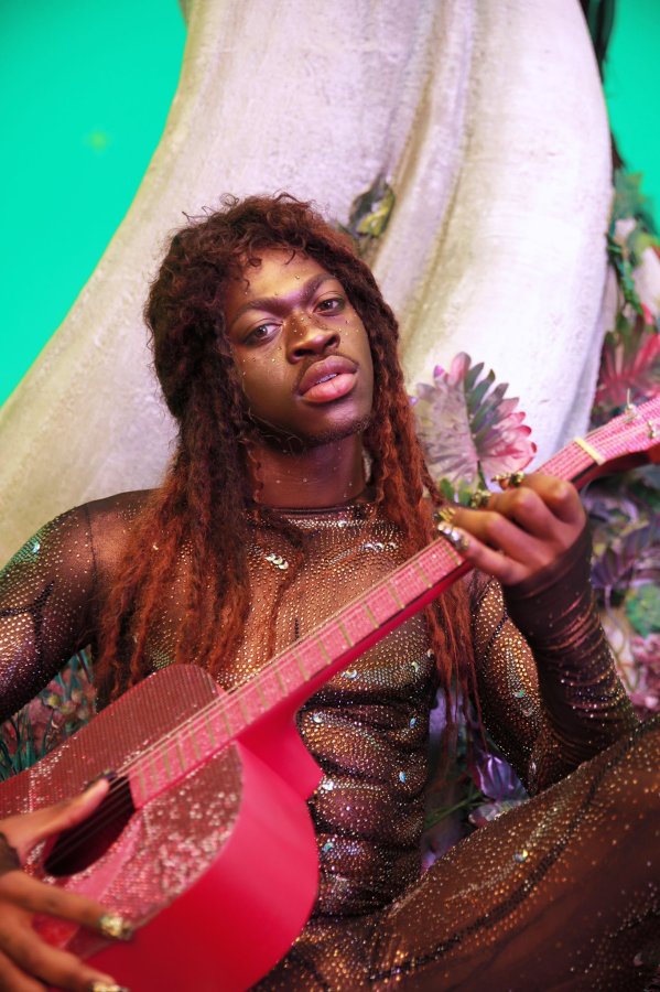 Image shows Lil Nas X stroking a glitter guitar with wavy long locs and a black glitter body suit.