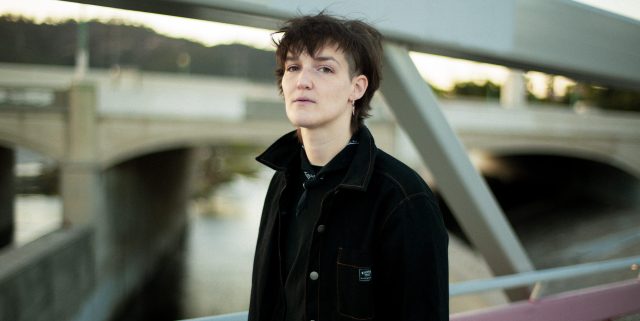 Artist Grace Semler Baldridge poses on a bridge. They are wearing a black coat and have a neutral expression.