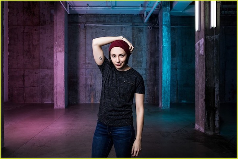 A close up of the actor Lauren Patten in a Burgundy red beanie cap and a dark grey t shirt. Her hand is over her head and she stares directly into the camera.