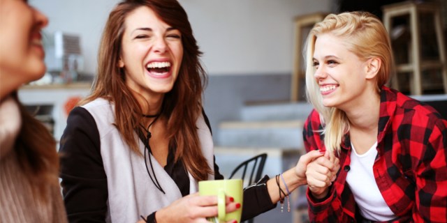 Two lesbians hold hands while laughing and sitting down. One lesbian is a brunette with a green mug and a grey shirt, one is a blonde with a flannel shirt.
