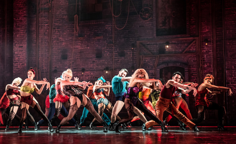 A still from the play Moulin Rouge, where the cast is in various fancy french lingerie and are dancing in front of a plain brick wall