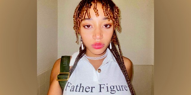 Actor Amandla Stenberg is in a white tank top that has "Father Figure" written in black letters. They have pink lip gloss and curly hair tendrils around their face.