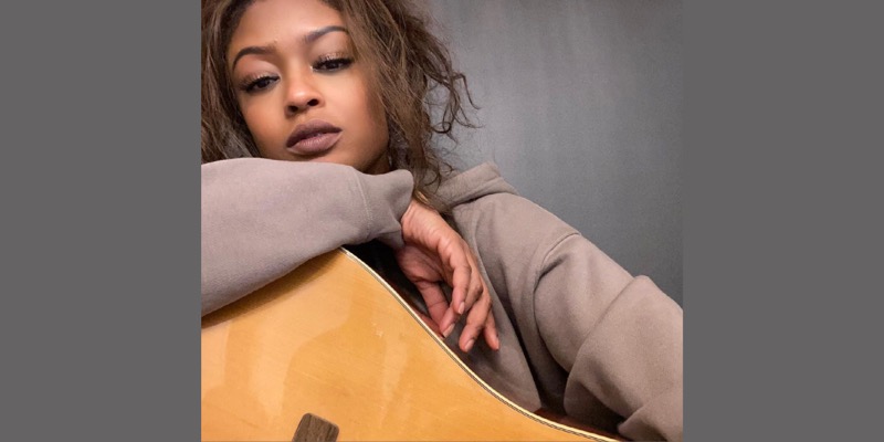 Actress Javicia Leslie is in a taupe brown sweatshirt with her her hair in a loose curly ponytail. She is holding a guitar and looking at the camera with her eyes low.