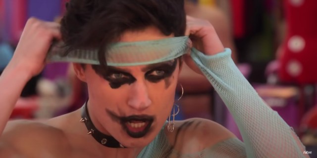 A contestant on RuPaul's Drag Race has smudged make up dark black lipstick and heavy eyeliner, they are putting on an aquamarine net headband