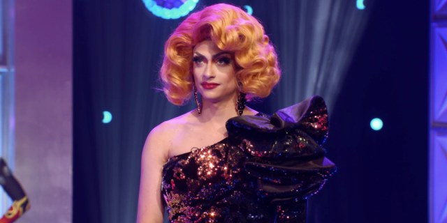 Denali is in an orange wig and a one shoulder sparkly brown dress with a large brown bow. She is on the runway of RuPaul's Drag Race with sparkly purple lights behind her.