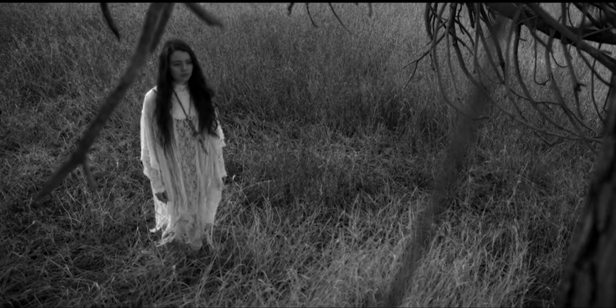 A still of a moment from Jayli Wolf's "Child of the Government" video, in which the artist stands facing a tree in a field wearing a flowing white nightgown.