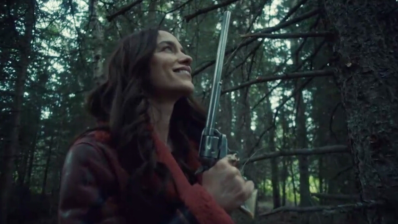 Wynonna Earp stands in the woods, smiling a sultry smile at Peacemaker, her trusty buntline special.