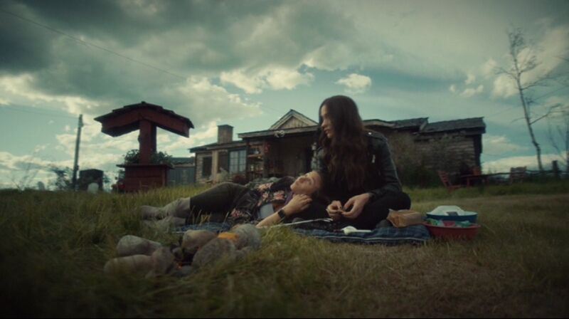 The Earp sisters sit by a fire, Waverly lying down with her head resting in Wynonna's lap.