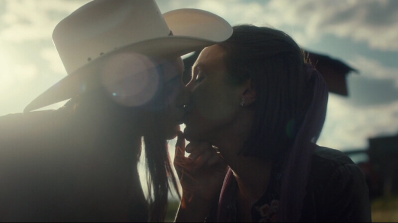 Nicole in her stetson kisses Waverly while gently lifting her chin, the light flaring softly around them.