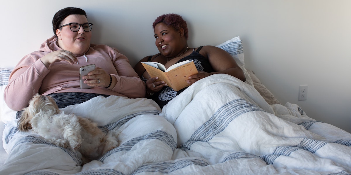 Two people curled up in bed lean into each other smiling as one reads a book and the other scrolls on their phone