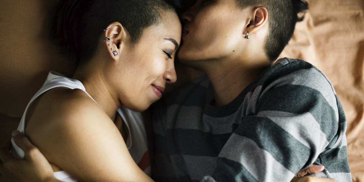 A person in a white tank top and a person in a striped shirt hold each other in bed. The person in the striped shirt kisses the other person's forehead.