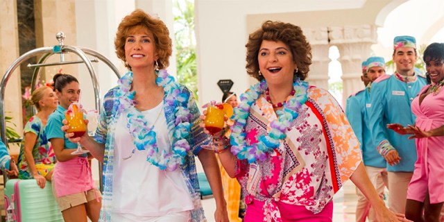 Annie Mumolo and Kristin Wiig walk through a bright hotel lobby holding frozen cocktails.