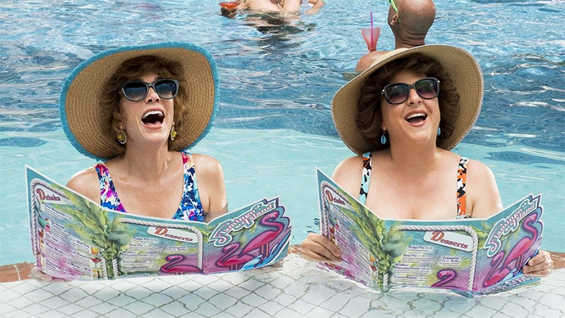 Annie Mumolo and Kristin Wiig wear giant visors in a hotel pool. 