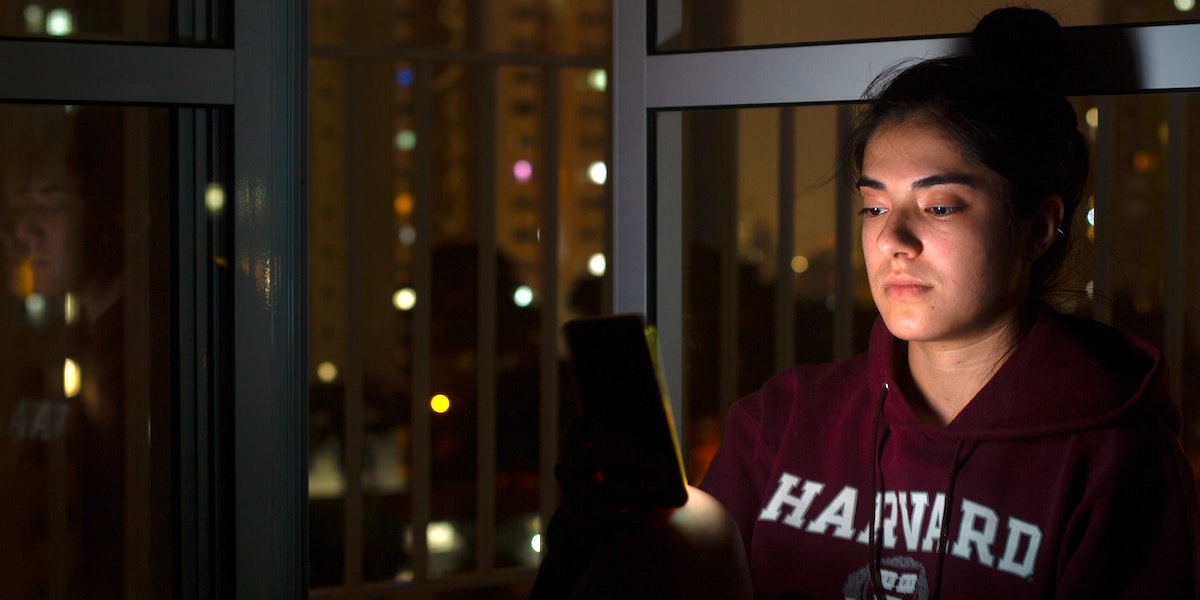 A young woman with her hair in a bun and wearing a college hoodie leans up against a window at night, staring at the screen of her smartphone with a serious expression