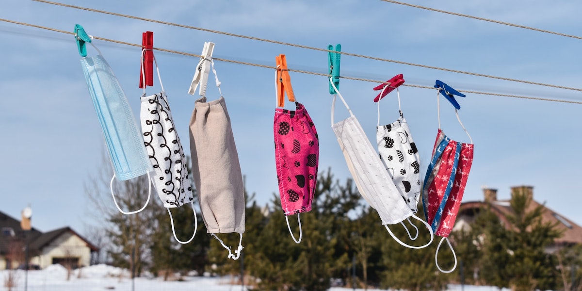 A photo of a series of colorful cloth face masks pinned to a clothesline against a background of trees in the distance and a blue sky