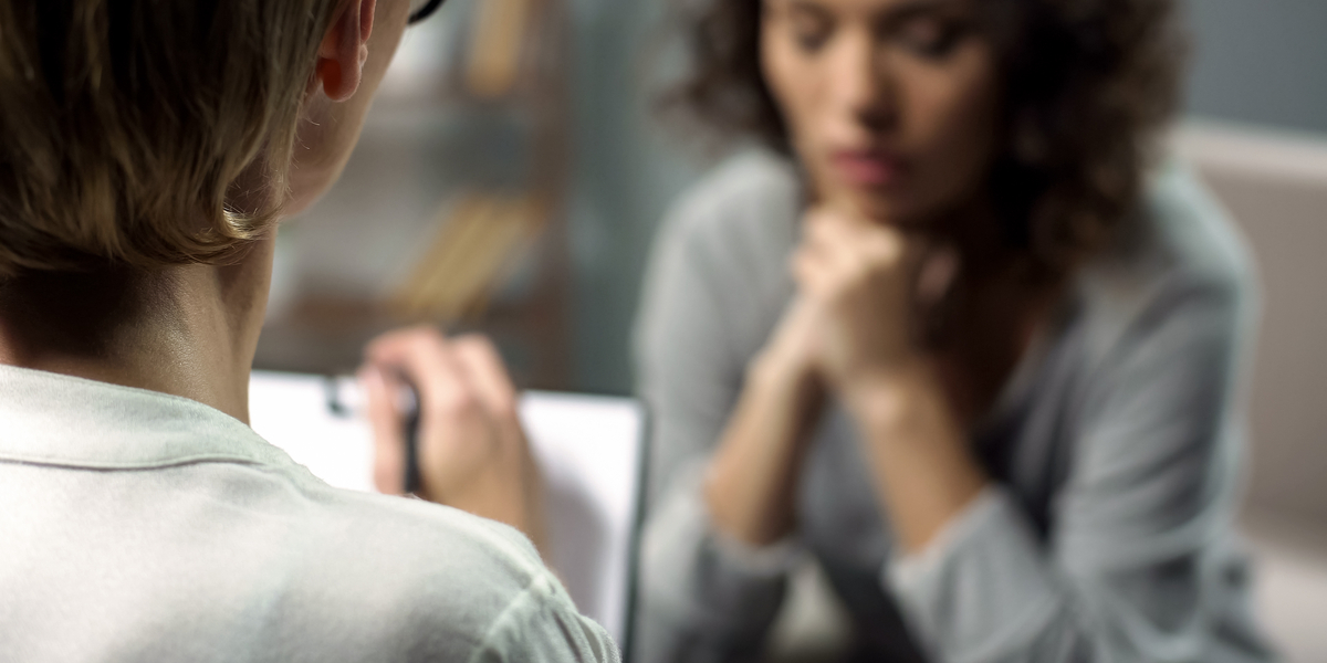 A young woman in soft focus in the background leans forward on her clasped hands speaking to a therapist with close-cropped hair in the foreground