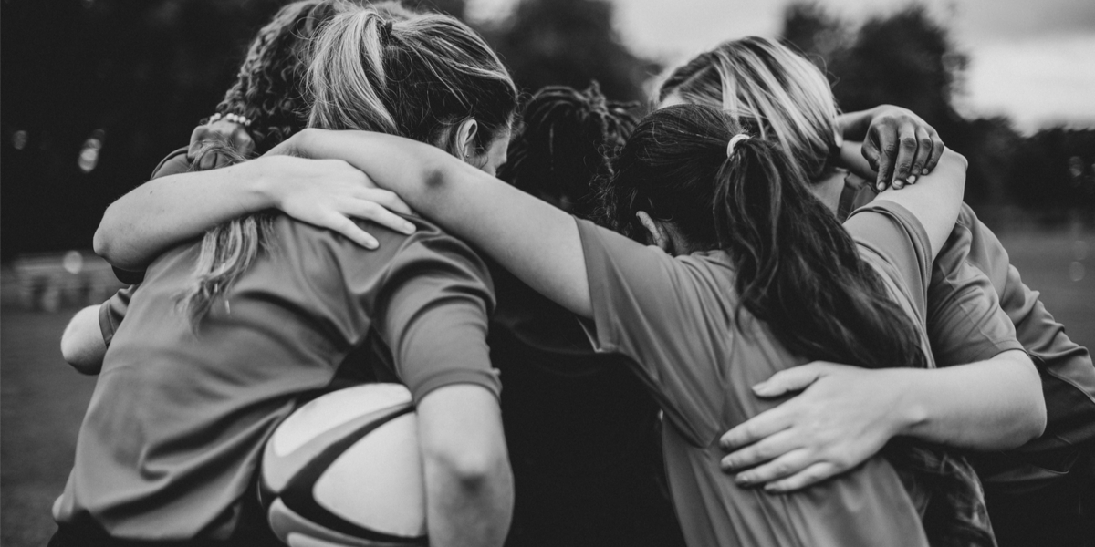 A black and white photo of teen rugby players huddled together, facing inwards in a cirlce and arms around each others' shoulders