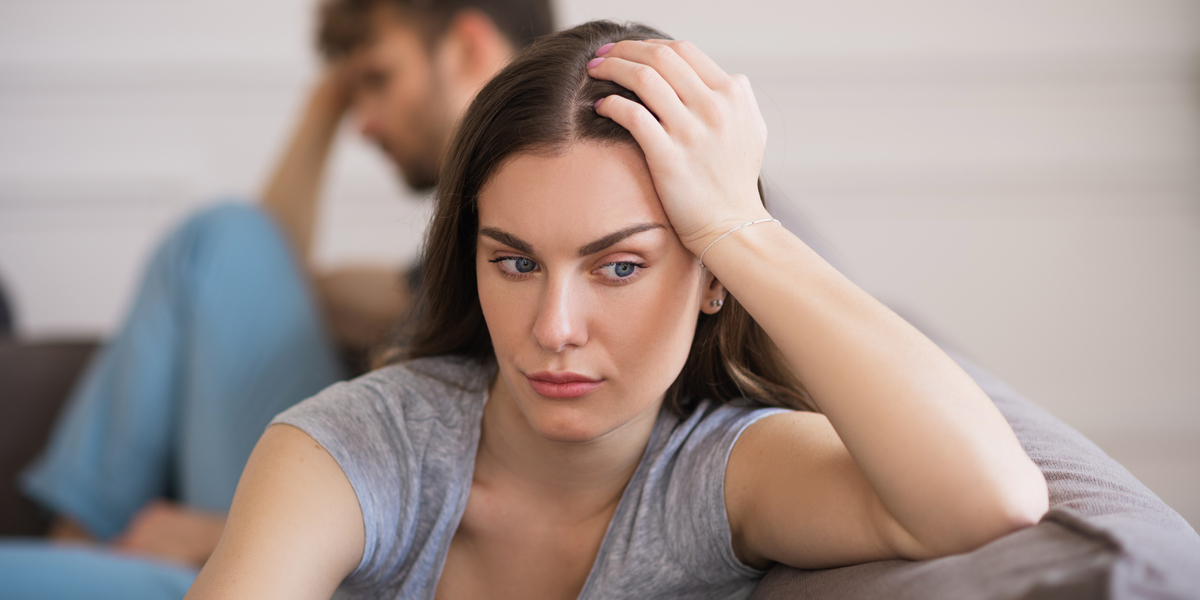 A photo of a man and woman sitting on a sofa; the woman looks off in a different direction as if lost in thought, a troubled expression on her face