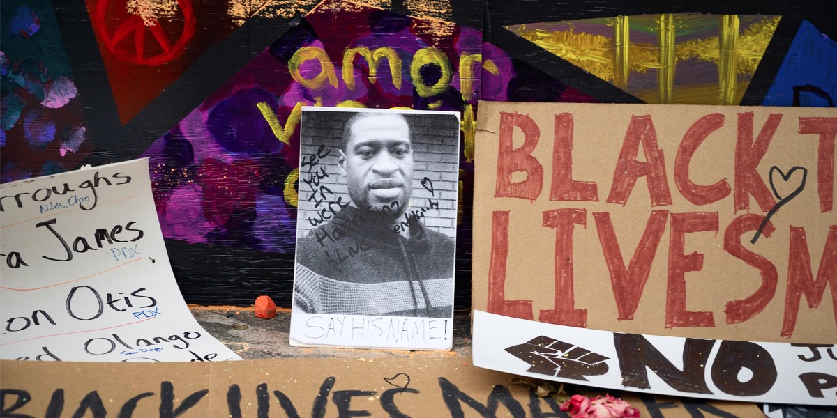 A protest sign featuring the face of George Floyd with the inscription "See You in Heaven" is laid on the ground next to a cardboard sign that reads "Black Lives Matter" in red sharpie