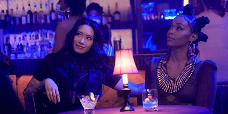 Grace and Anissa sit together at a table in sexy blue lighting.