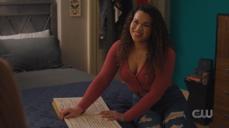 Josefina from Charmed sits on a bed with the Book of Shadows and smiles up at her cousin off screen.